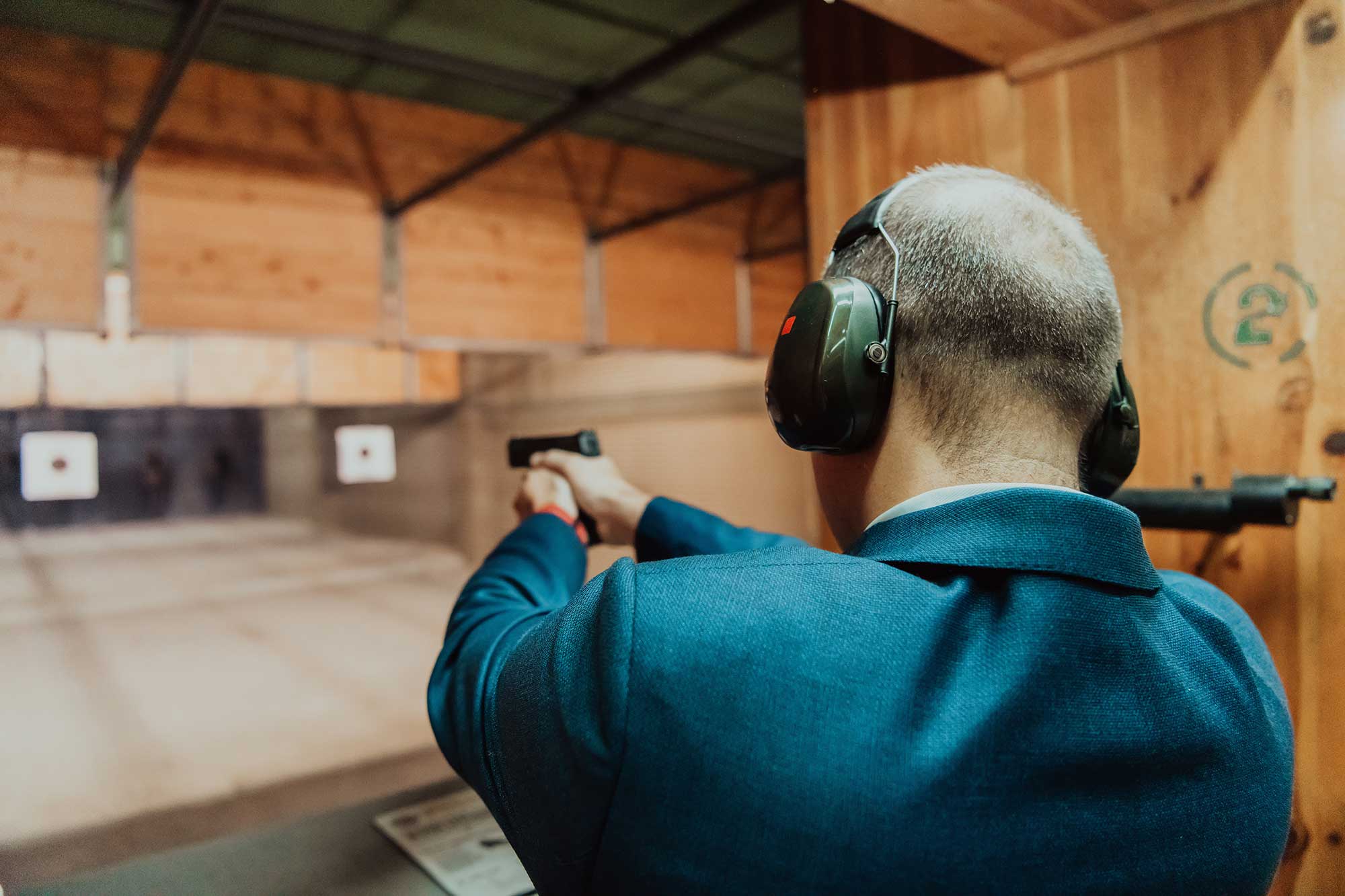 Man looking away from camera, aiming pistol at indoor firing range while wearing earmuffs.